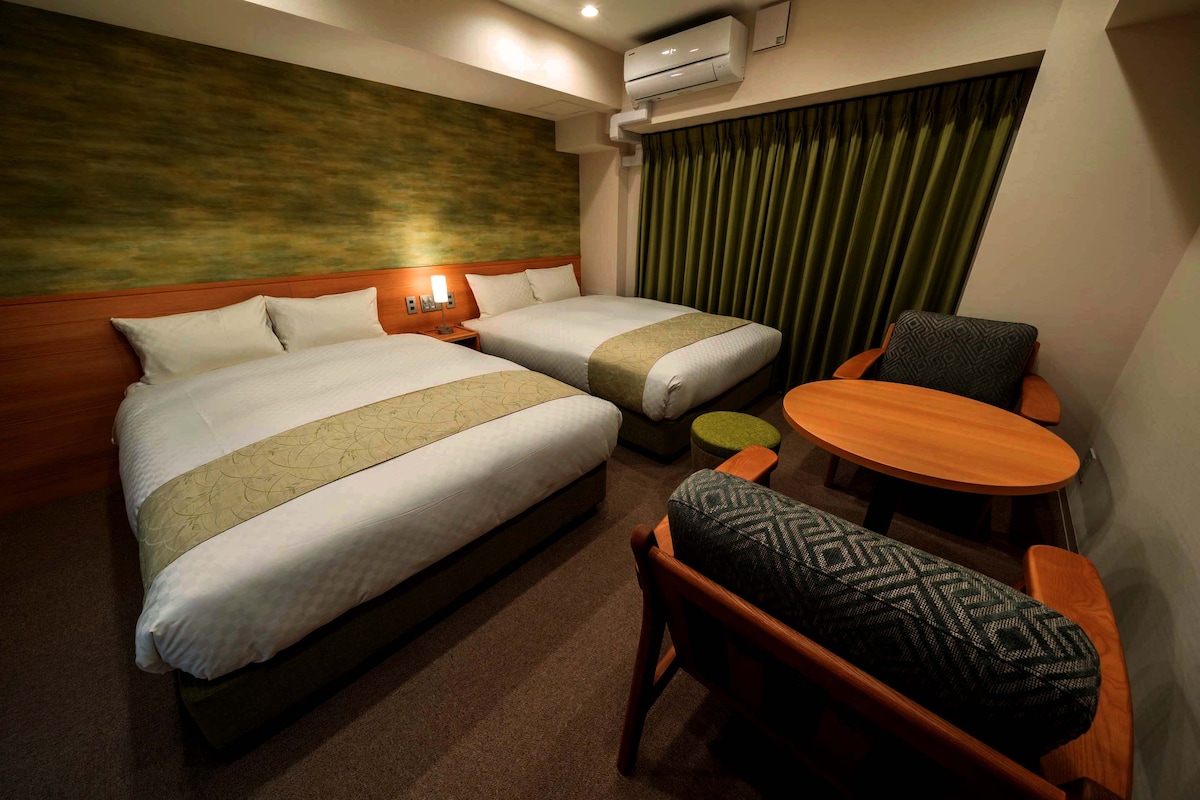 The room is equipped with two spacious beds dressed in crisp white linens and accented with green bed runners. A round wooden table is accompanied by a couple of armchairs, creating a cozy seating area. The walls feature a calming green design, complemented by elegant curtains.