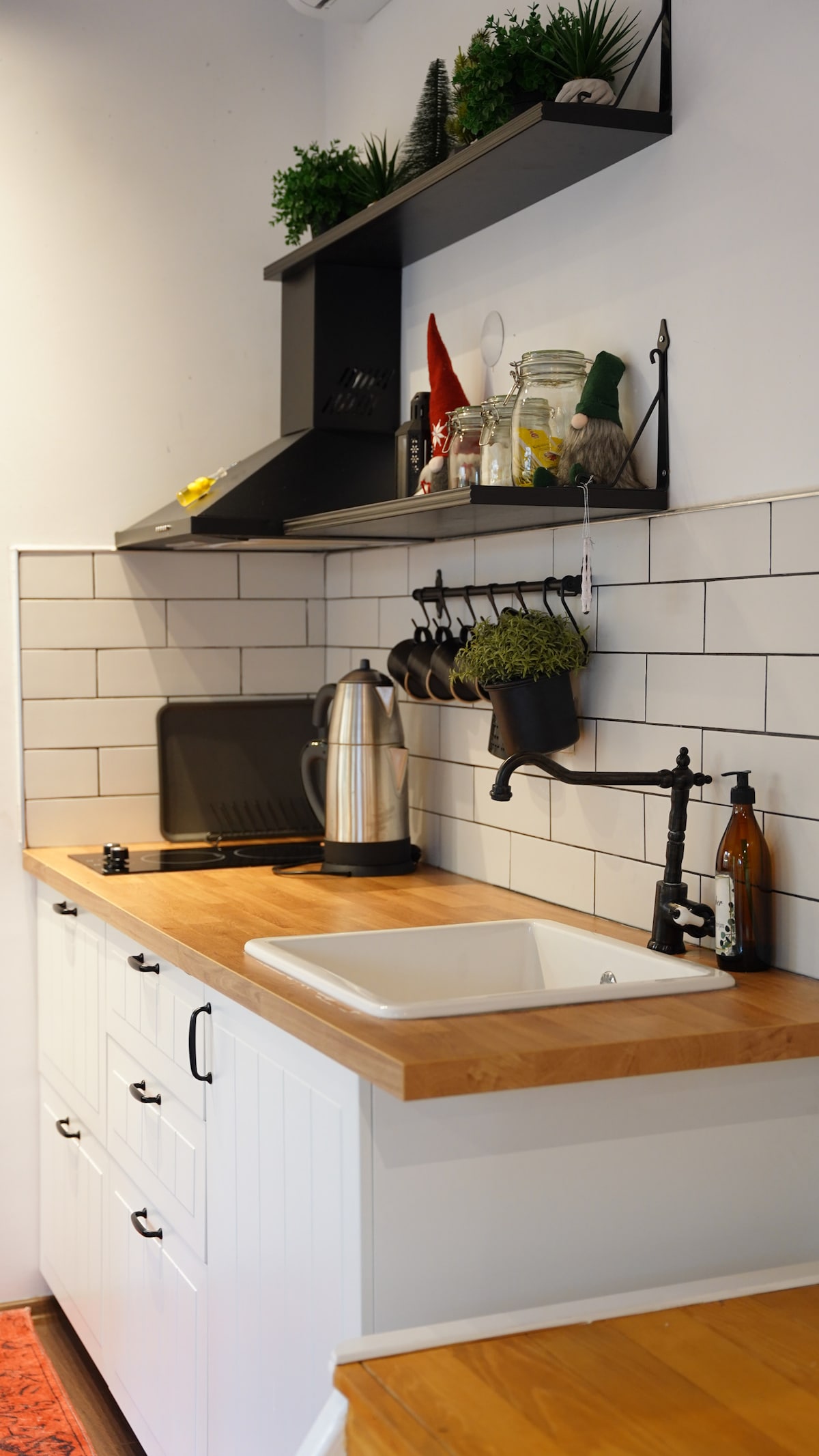 A modern kitchen features a wooden countertop with a white sink below an open shelf holding plants and cooking essentials. A kettle and black utensils are neatly arranged, with a black dish rack positioned beside the sink. Light tiles create a clean backdrop.