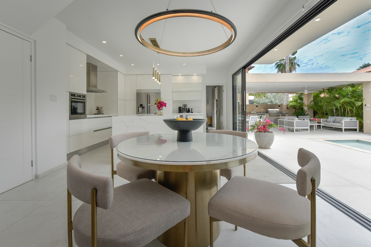 A modern dining area features a round table surrounded by four upholstered chairs. A pendant light hangs above the table, while expansive glass doors open to the outdoor space, revealing views of lush greenery and the pool outside.