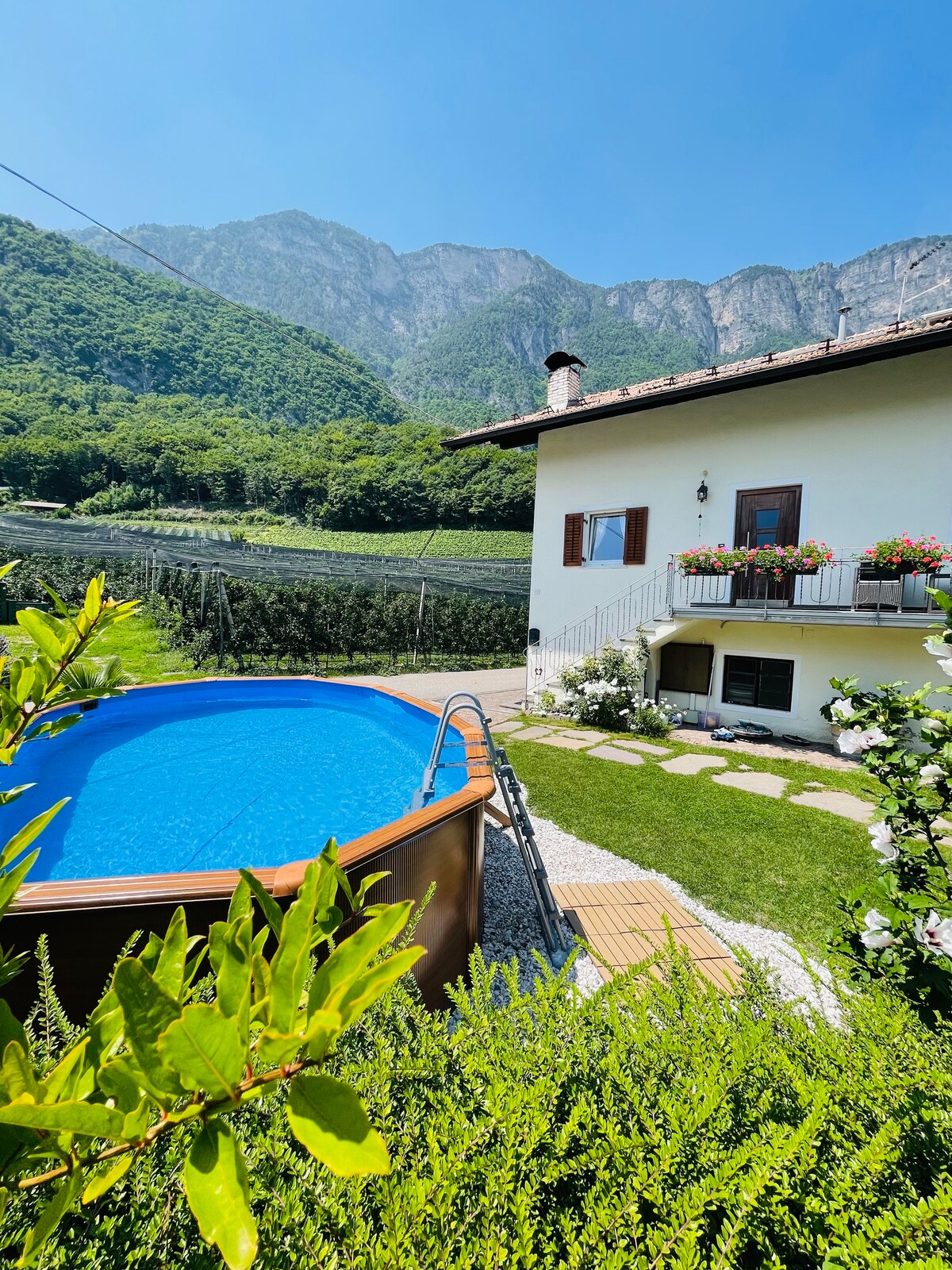 A spacious garden area features a round above-ground pool surrounded by shrubbery and flowers. The two-story building nearby displays wooden shutters and a balcony adorned with colorful plants, set against a backdrop of lush mountains.