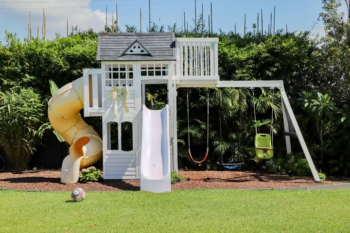 A children's white playset in a backyard, featuring a yellow spiral slide, a white straight slide, and swings. The structure is set against a backdrop of tall greenery with a soccer ball on the grassy lawn in front