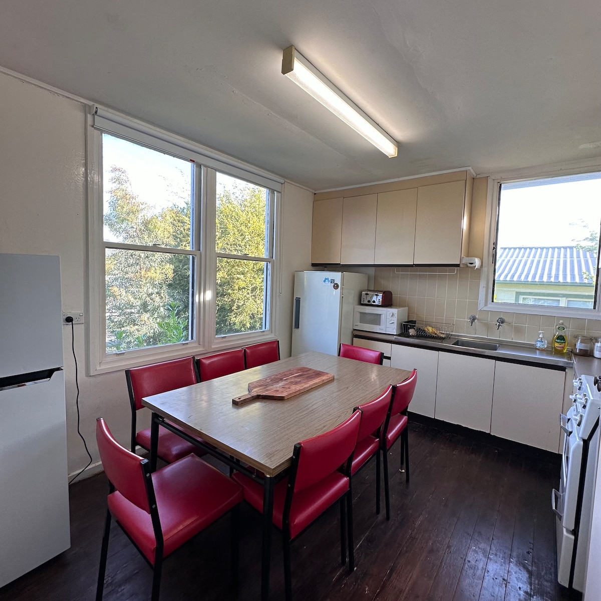 A functional kitchen features a large table surrounded by six red chairs. Natural light enters through two windows, illuminating the white cabinetry and appliances, including a refrigerator and microwave. A wooden cutting board rests on the table, adding a practical touch to the space.