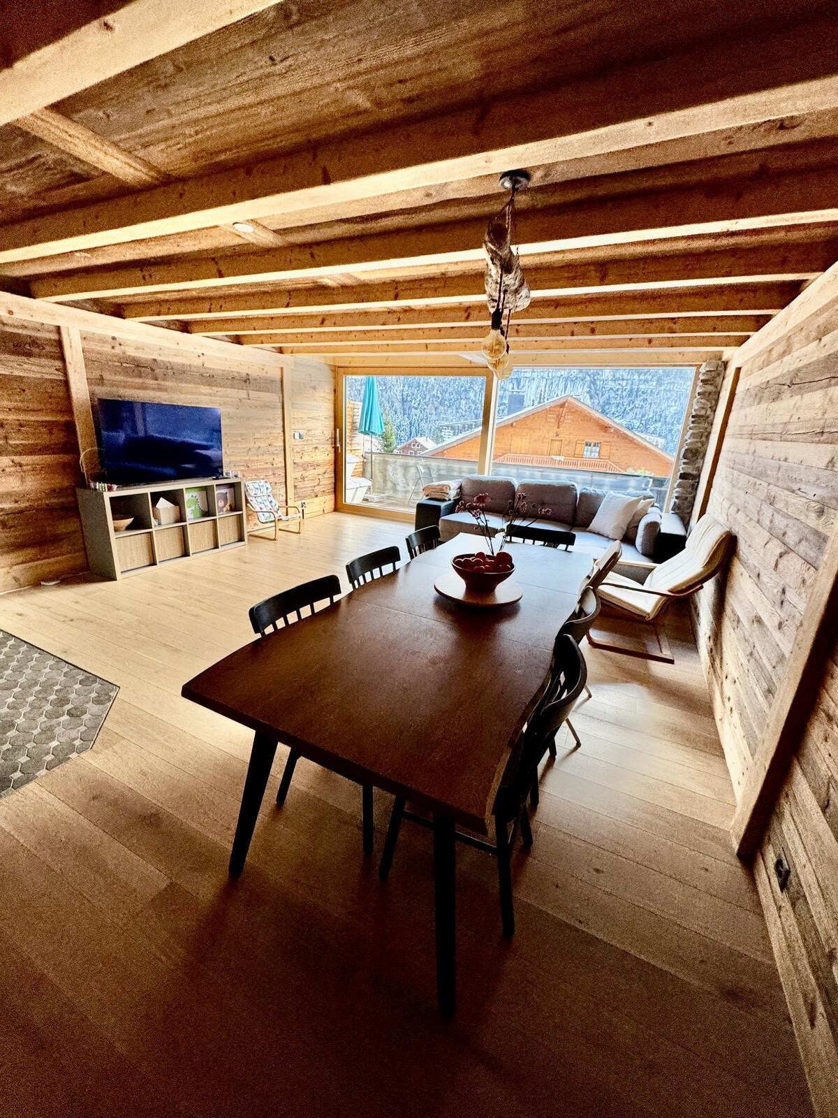 A spacious open-plan living area is characterized by warm wooden walls and ceiling beams. A large dining table with black chairs is positioned centrally, while a comfortable seating area with a television is visible beside large windows that overlook the outdoor scenery.