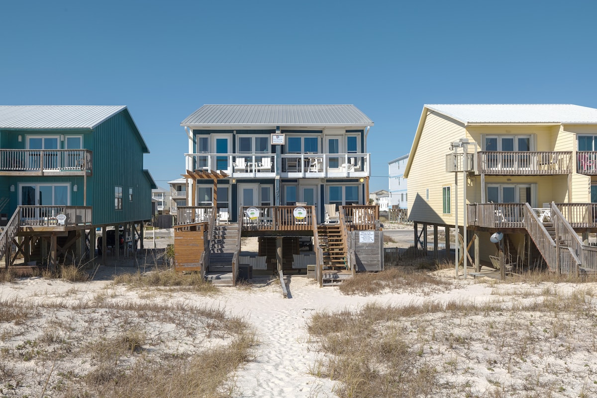 The beachfront duplex features a light exterior with multiple balconies overlooking the sandy pathway leading to the beach. Surrounding structures exhibit a colorful coastal aesthetic. The sandy area showcases minimal vegetation, providing a clear view of this serene coastal retreat.