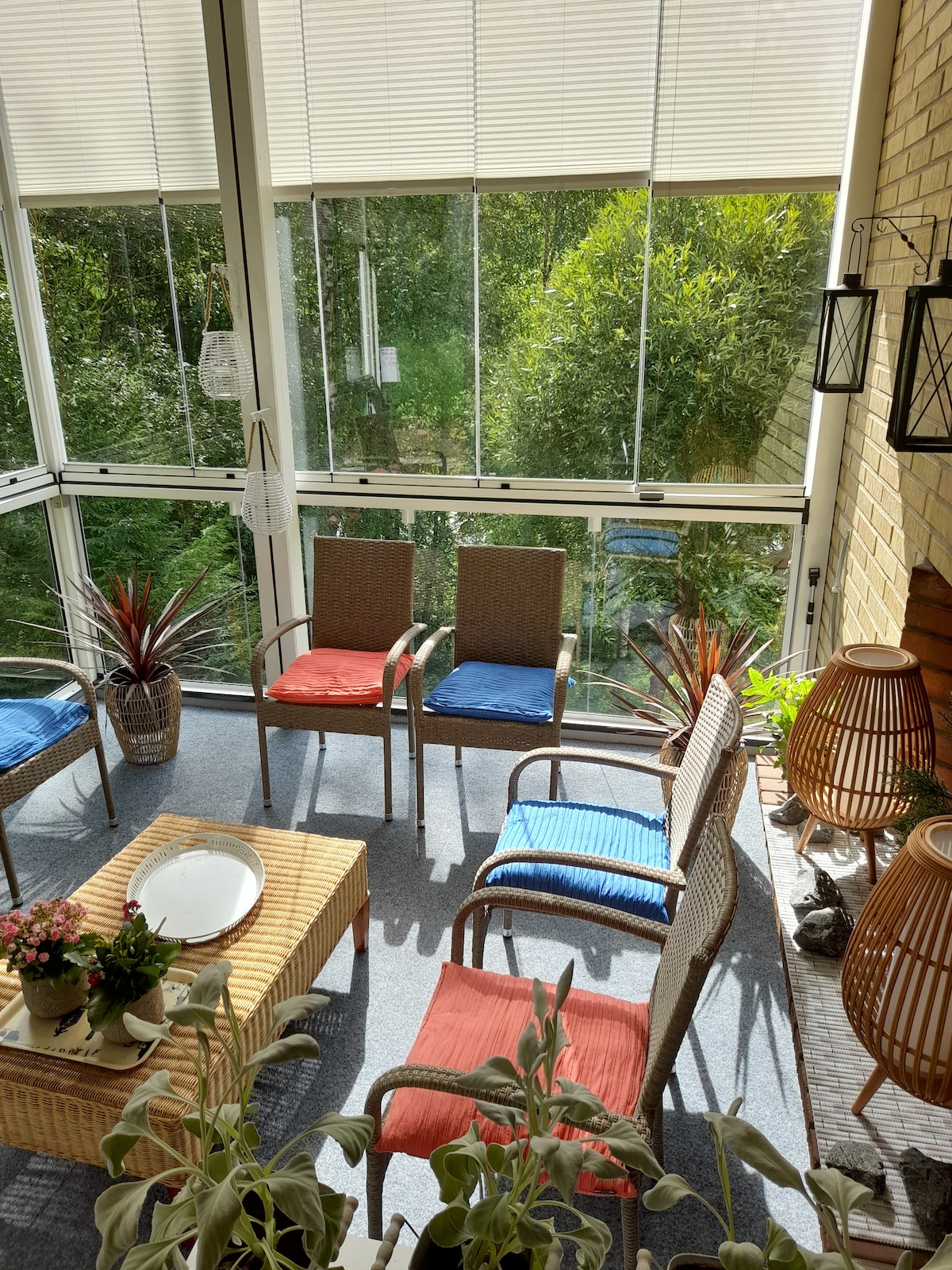 A sunlit indoor space features several chairs with colorful cushions arranged around a woven coffee table. Lush greenery can be seen through large glass windows, while decorative plants and lanterns add a touch of warmth to the atmosphere.