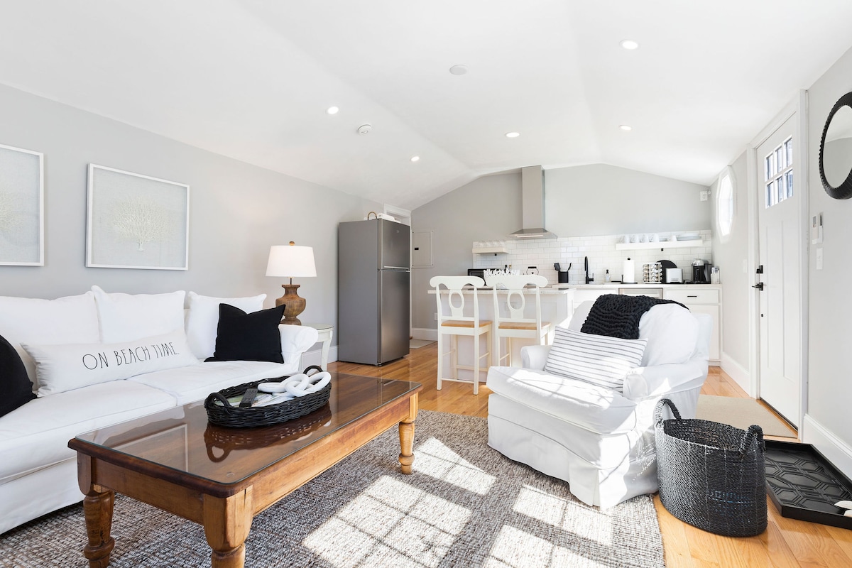 An open concept living area features a comfortable white couch and armchair, with a wooden coffee table at the center. The kitchen, visible in the background, is well-equipped and includes a fridge, while a dining area with two stools is highlighted. Natural light brightens the space.