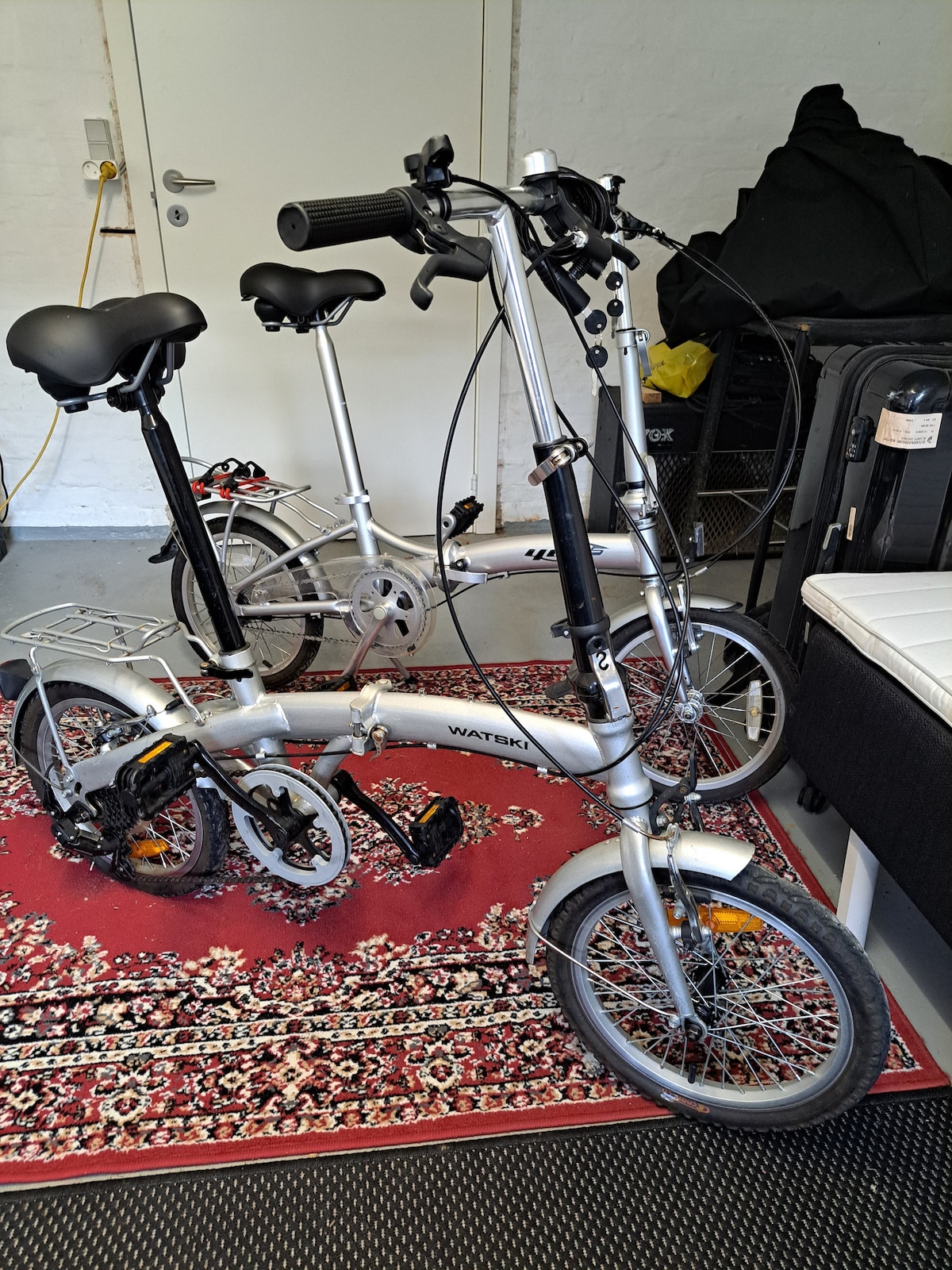 Two compact bicycles are positioned on a decorative area rug, showcasing a sleek silver design. The bicycles feature adjustable seats and handlebars, with visible gear mechanisms and pedals. A black suitcase and additional items are subtly included in the background.