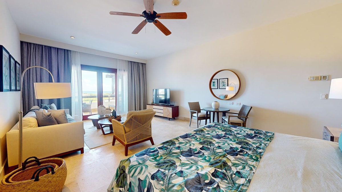 A spacious studio room features a king-sized bed dressed with a patterned duvet. A modern seating area includes a sofa and two chairs, with a round dining table nearby. Natural light fills the space through large sliding doors, leading to a balcony with scenic views.