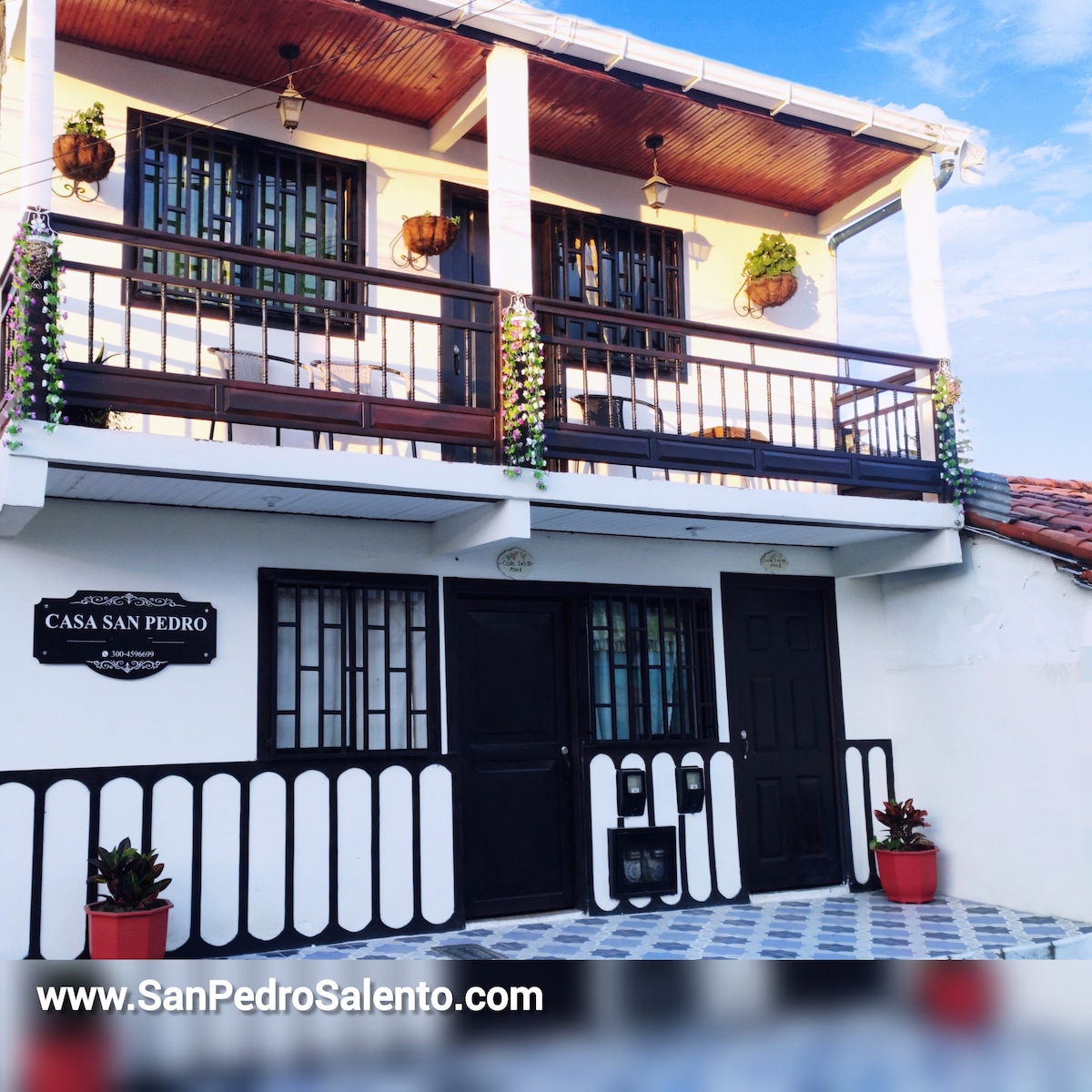 The exterior of the apartment building is featured, showcasing a white facade with dark wooden accents. A welcoming entrance leads to a covered porch adorned with potted plants. Large windows and a balcony above create an airy atmosphere, complemented by patterned floor tiles.