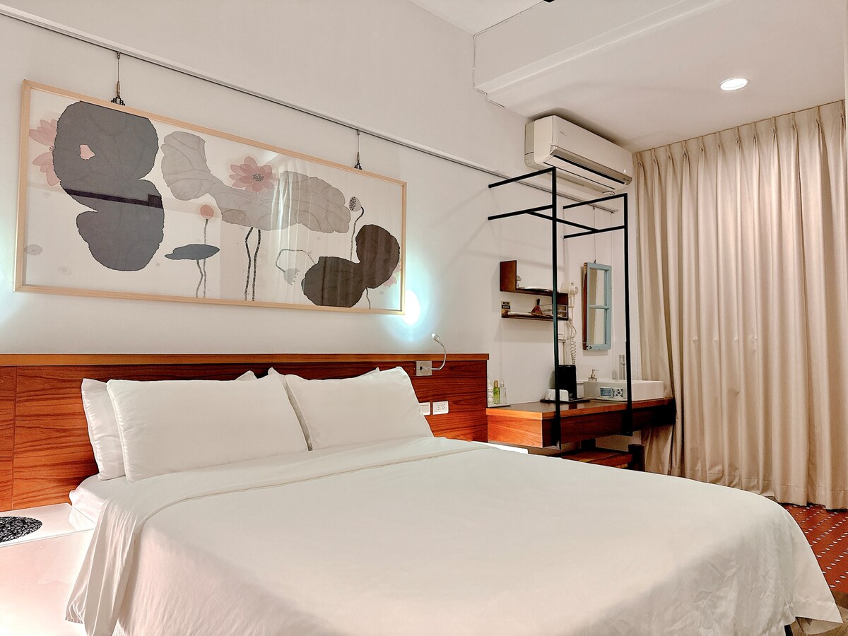 A spacious sleeping area is highlighted by a standard comfortable double bed, adorned with crisp white bedding. A large artwork decorates the wall above the bed, while a simple desk and mirror are positioned to the side. Soft curtains drape over the window, allowing gentle light in.