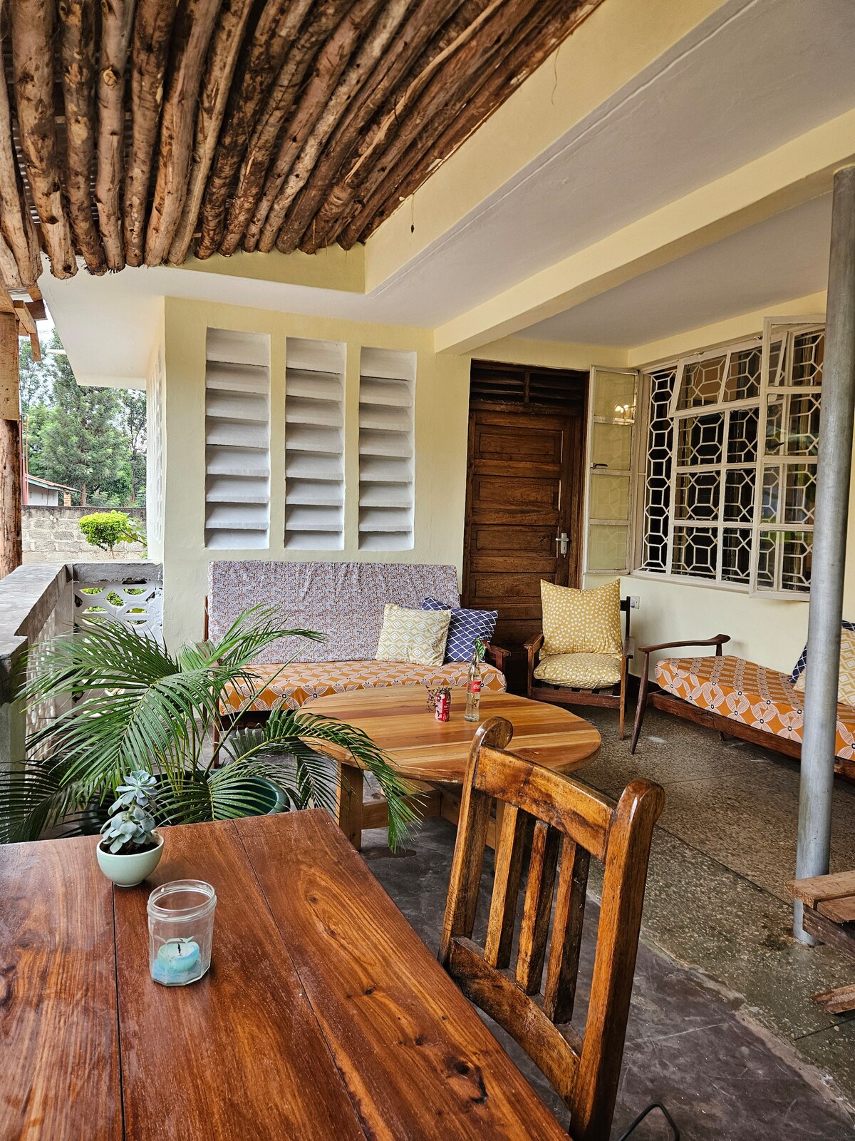 An inviting outdoor seating area features a wooden table accompanied by several chairs. Cushions in vibrant patterns adorn a comfortable sofa, complemented by potted plants. Natural light flows from nearby windows, while decorative wooden beams grace the ceiling, creating a tranquil space for relaxation.