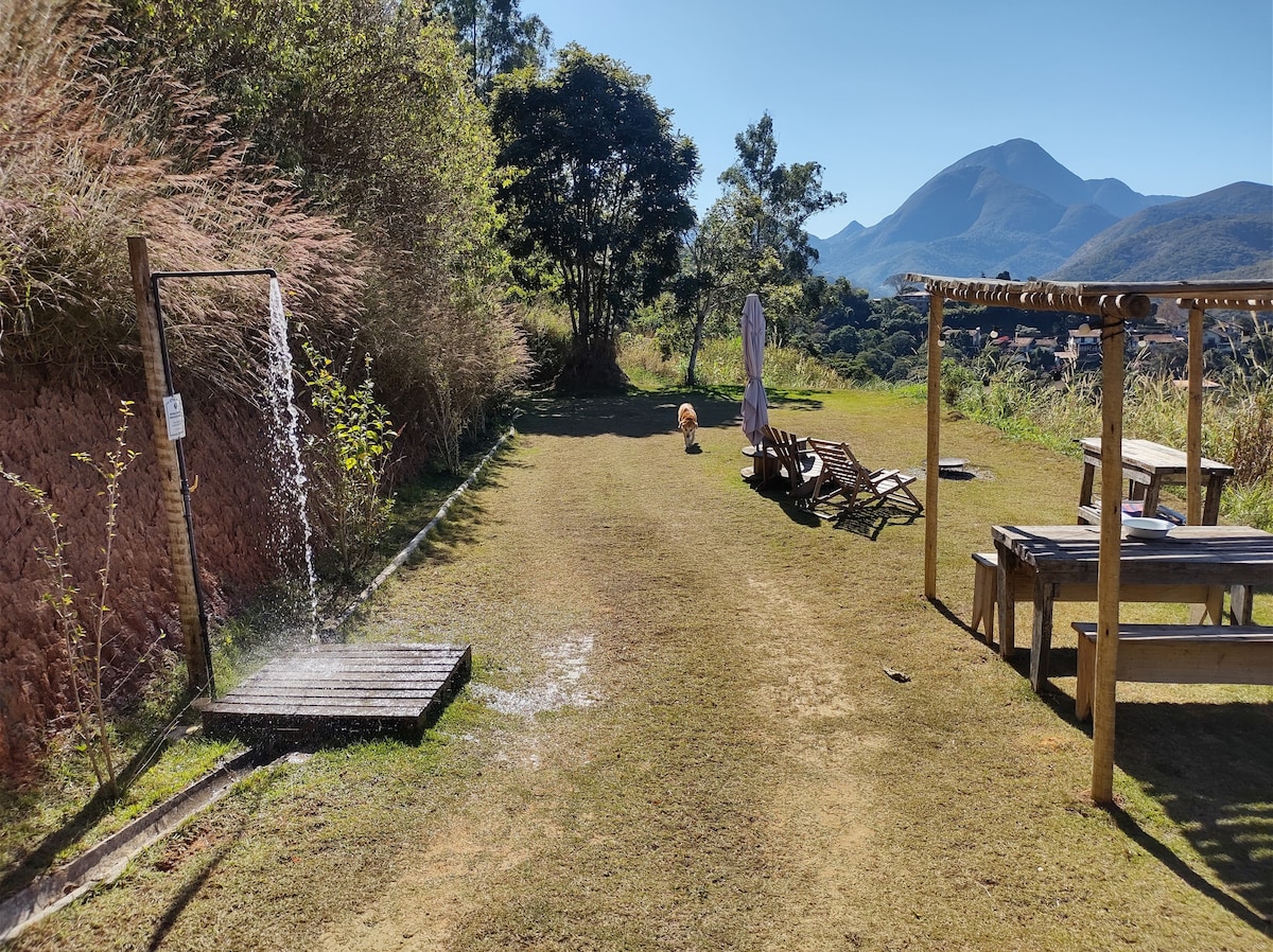 An outdoor area is presented with a grassy space featuring a wooden shower, a sunshade, and rustic seating arrangements. Rolling hills and mountains are visible in the background, creating a serene landscape. A playful dog can be seen in the distance.