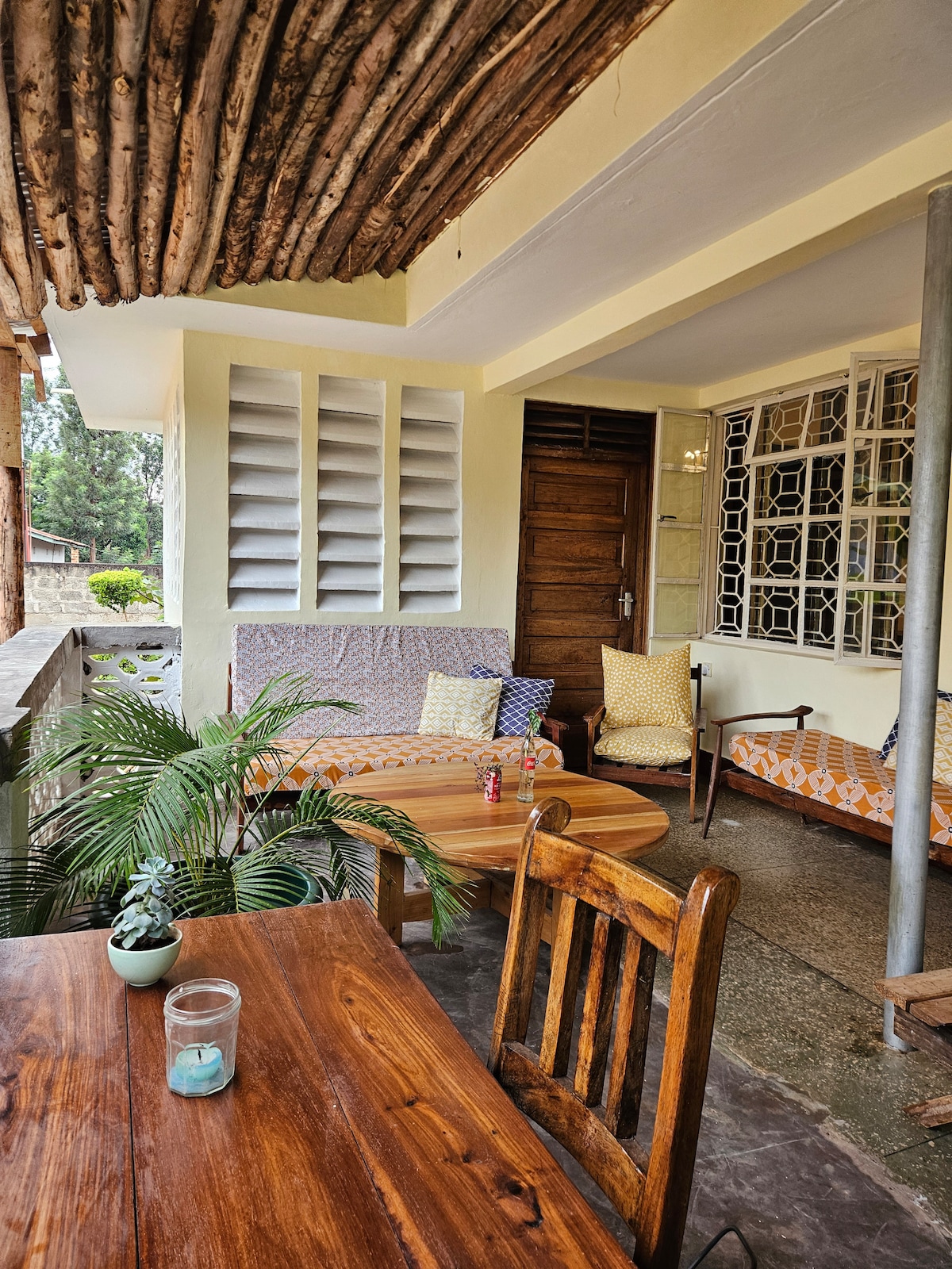 A shared outdoor seating area features comfortable cushions on wooden furniture. A small table with a glass and a plant can be seen, enhancing the space's welcoming feel. Wooden beams support the ceiling, while natural light streams through large windows.