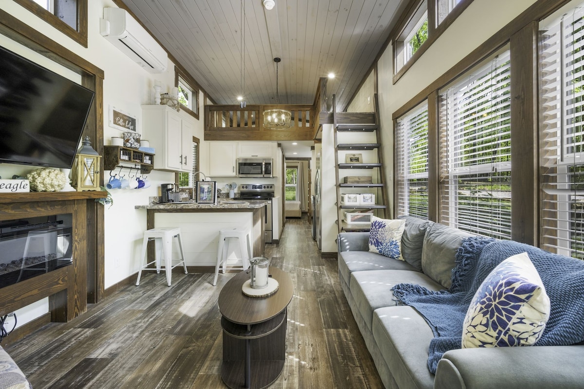 The living area features a spacious couch adorned with patterned pillows, arranged around a circular coffee table. A flat-screen TV is mounted on the wall, and a kitchen with modern appliances can be seen in the background. Large windows allow natural light to fill the space.