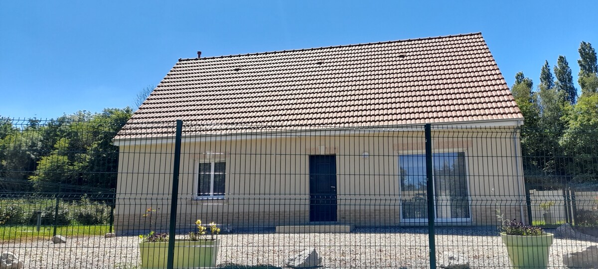 A single-story house is framed by a green garden, with a fenced yard visible in the foreground. The exterior features light-colored walls and a well-maintained, sloped roof. A central entrance leads to a spacious window on one side, while a smaller window is positioned to the left.