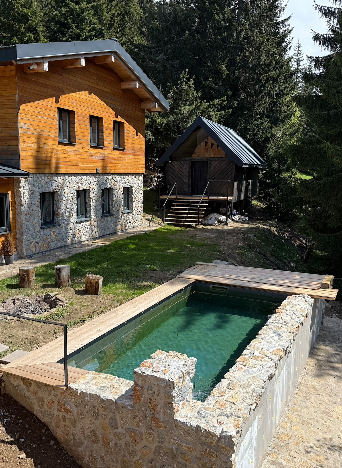 An outdoor swimming pool is surrounded by natural stone edges and wooden decking. The pool reflects the surrounding greenery, with trees visible in the background. A wooden cabin is positioned nearby, accompanied by stone seating arranged outdoors.