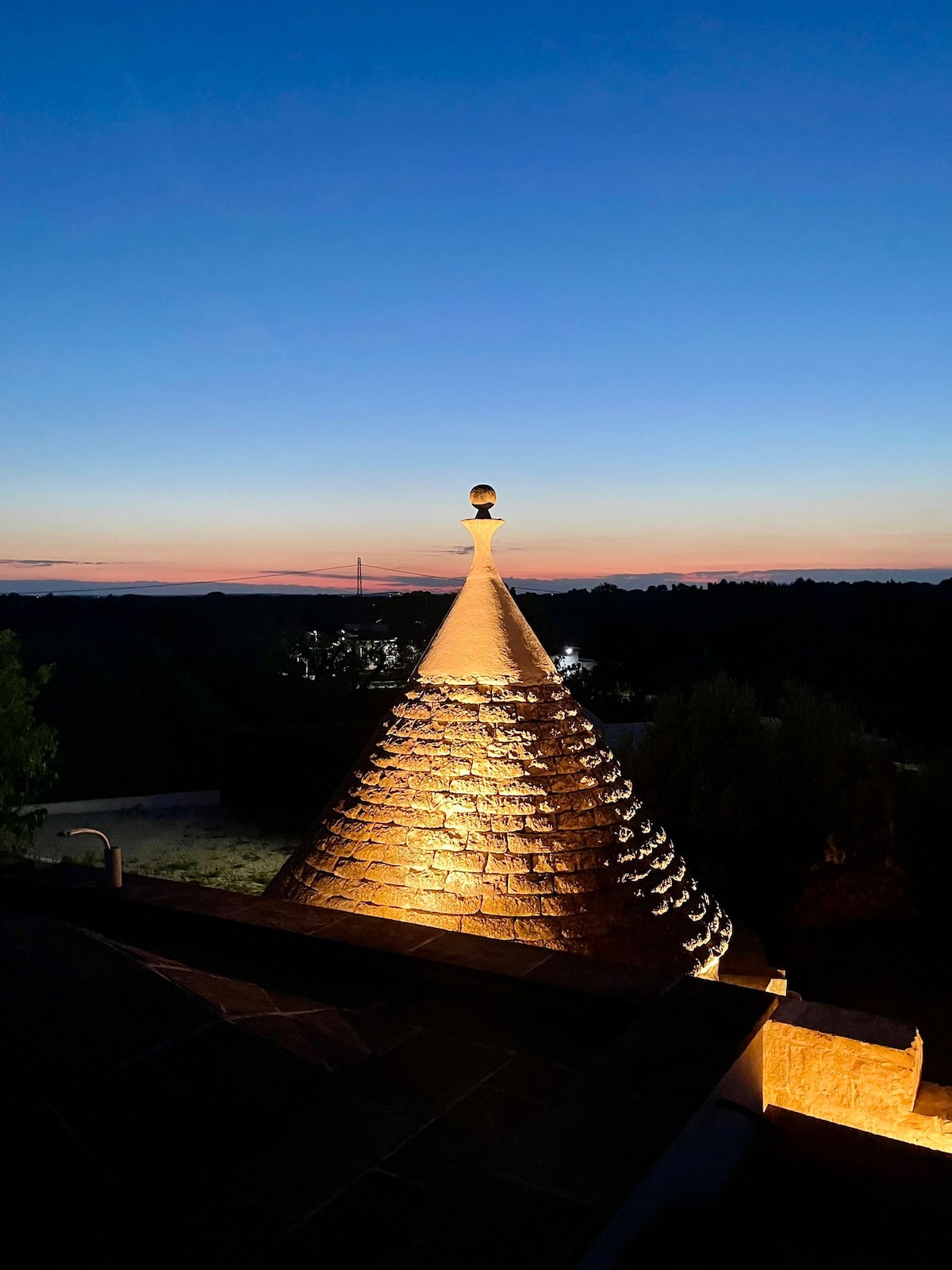 The image captures a conical rooftop structure illuminated by soft lighting, showcasing textured stonework. A gradient sky extends into twilight hues of blue and orange, while distant outlines of trees and landscape can be seen under the fading light.