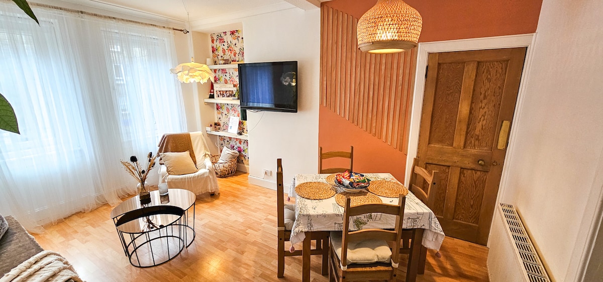 A cozy living area showcases wooden flooring and a soft seating space. A circular coffee table is placed in front of a small television mounted on the wall. A simple dining table with placemats is set up, adjacent to a sunlit area with decorated walls.
