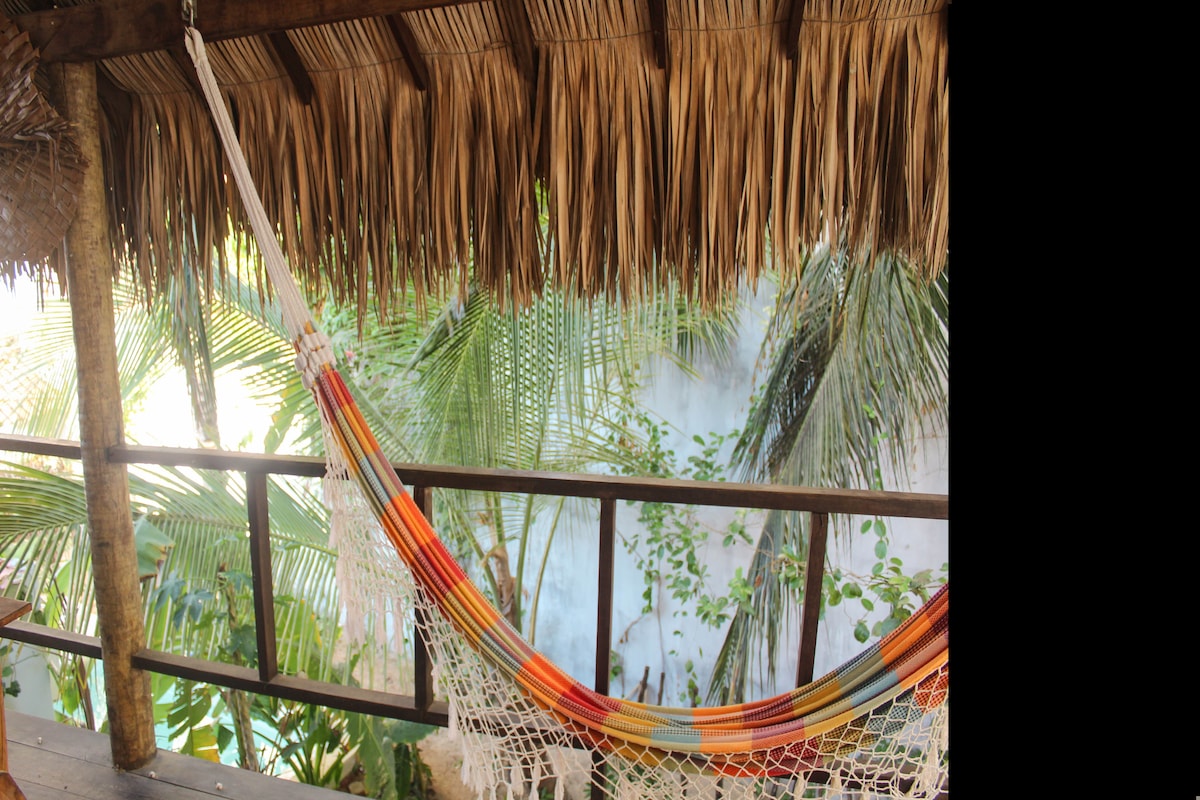A colorful hammock is suspended under a thatched roof, surrounded by lush tropical foliage. Sunlight filters through the leaves, creating a serene setting for relaxation. The wooden railing complements the natural decor, enhancing the peaceful ambiance of the space.
