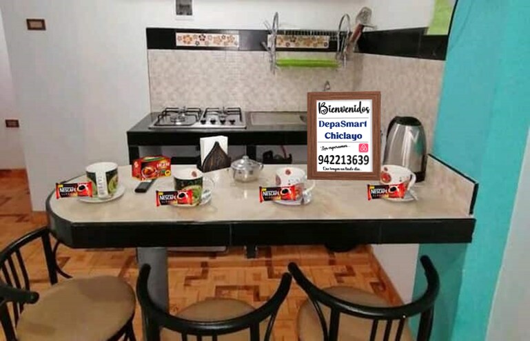 A compact kitchen area features a countertop with several bowls and jars of instant coffee. A kettle is seen alongside a gas stove against a backdrop of light cabinetry and tiled details. Two chairs are positioned at the countertop, contributing to a functional space.