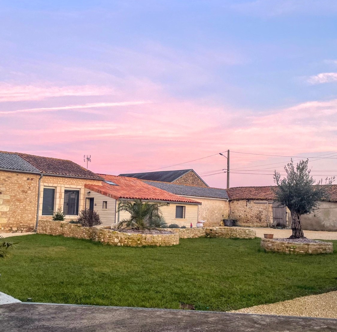 A serene outdoor space features a well-maintained lawn surrounded by stone walls and historic buildings. The sky displays soft hues of pink and blue, while a pathway leads into the grassy area, offering a tranquil setting for relaxation.