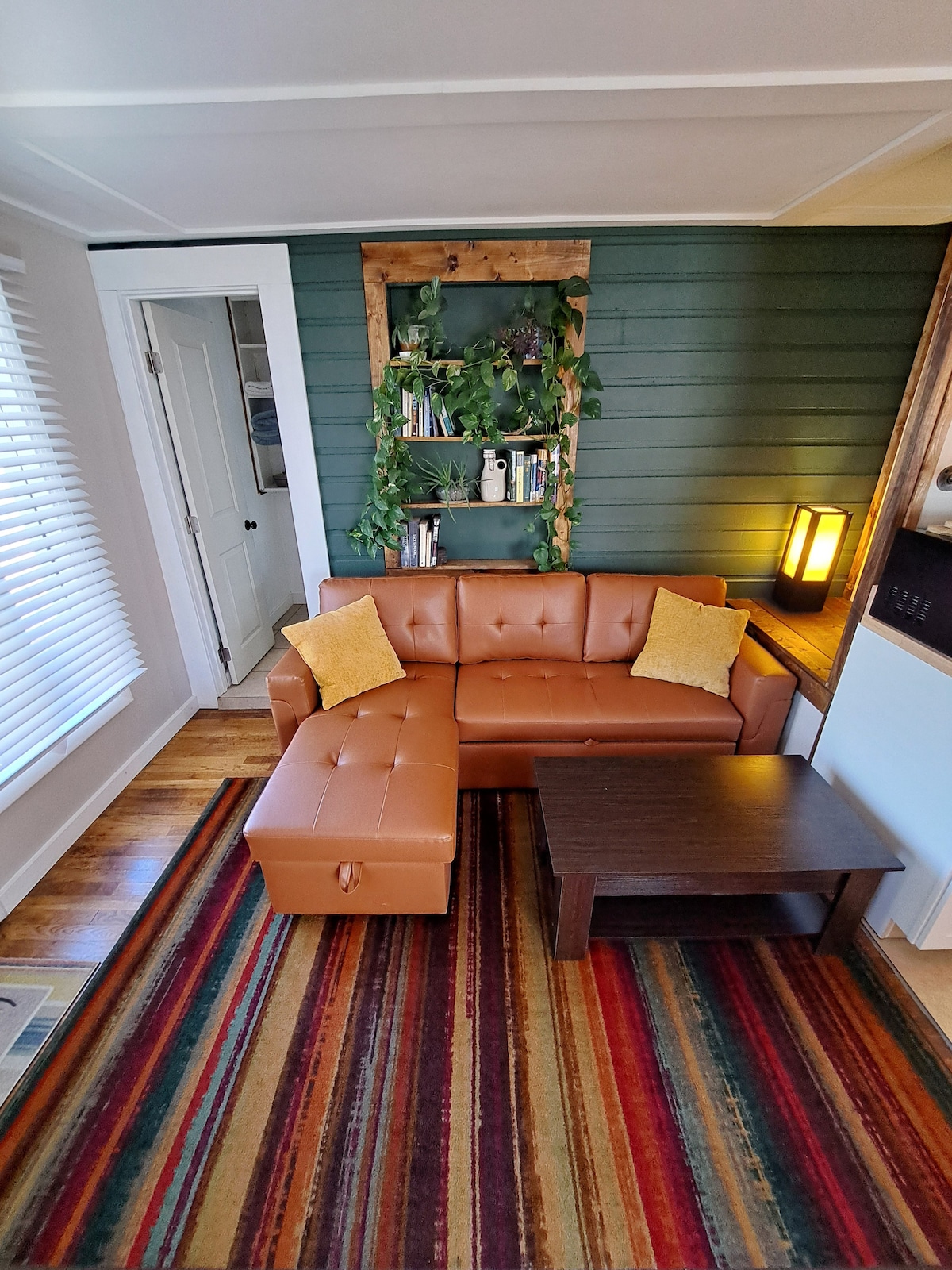 A cozy living area is shown, featuring a brown leather couch with yellow cushions arranged comfortably. A small coffee table sits in front on a striped area rug, and a wooden shelf with greenery can be seen against the green wall, adding to the inviting atmosphere.
