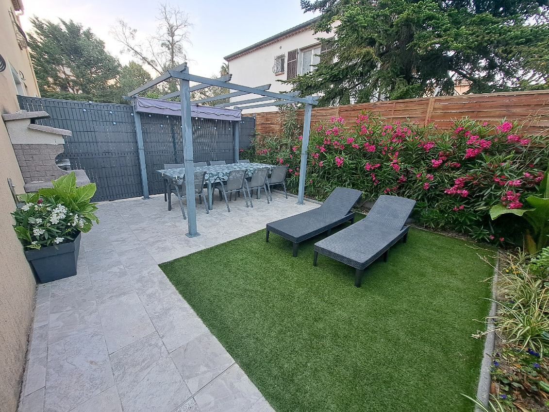 An outdoor space features a covered dining area with a large table and chairs. Two lounge chairs are positioned on a grassy area, surrounded by lush greenery and flowering plants. A stone patio complements the setting, offering a comfortable retreat.