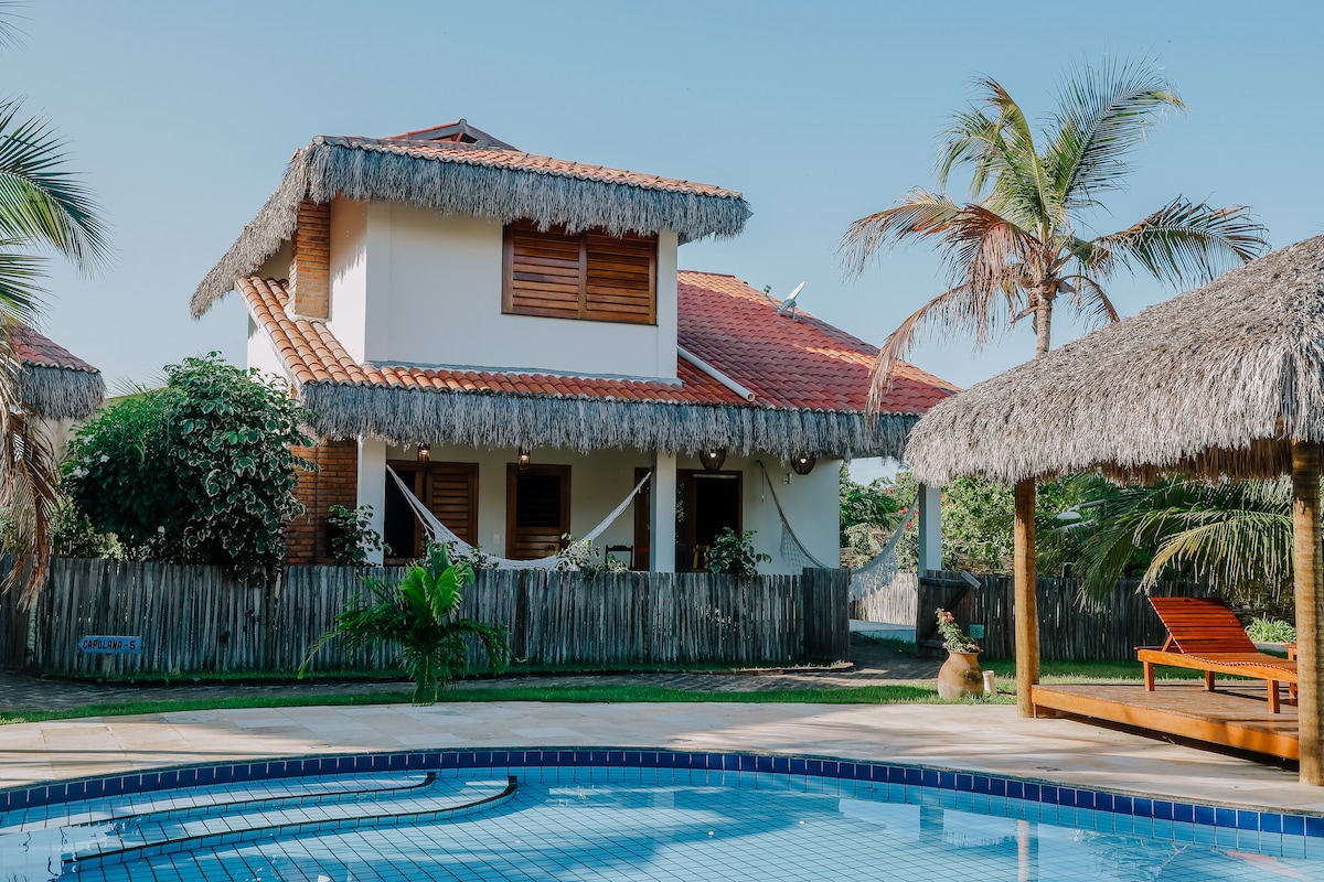 A two-story house features a thatched roof and wooden shutters, surrounded by lush greenery. A swimming pool is visible in the foreground, while a sunbed and shaded seating area enhance the outdoor setting. Tropical palm trees frame the scene, creating a relaxed atmosphere.