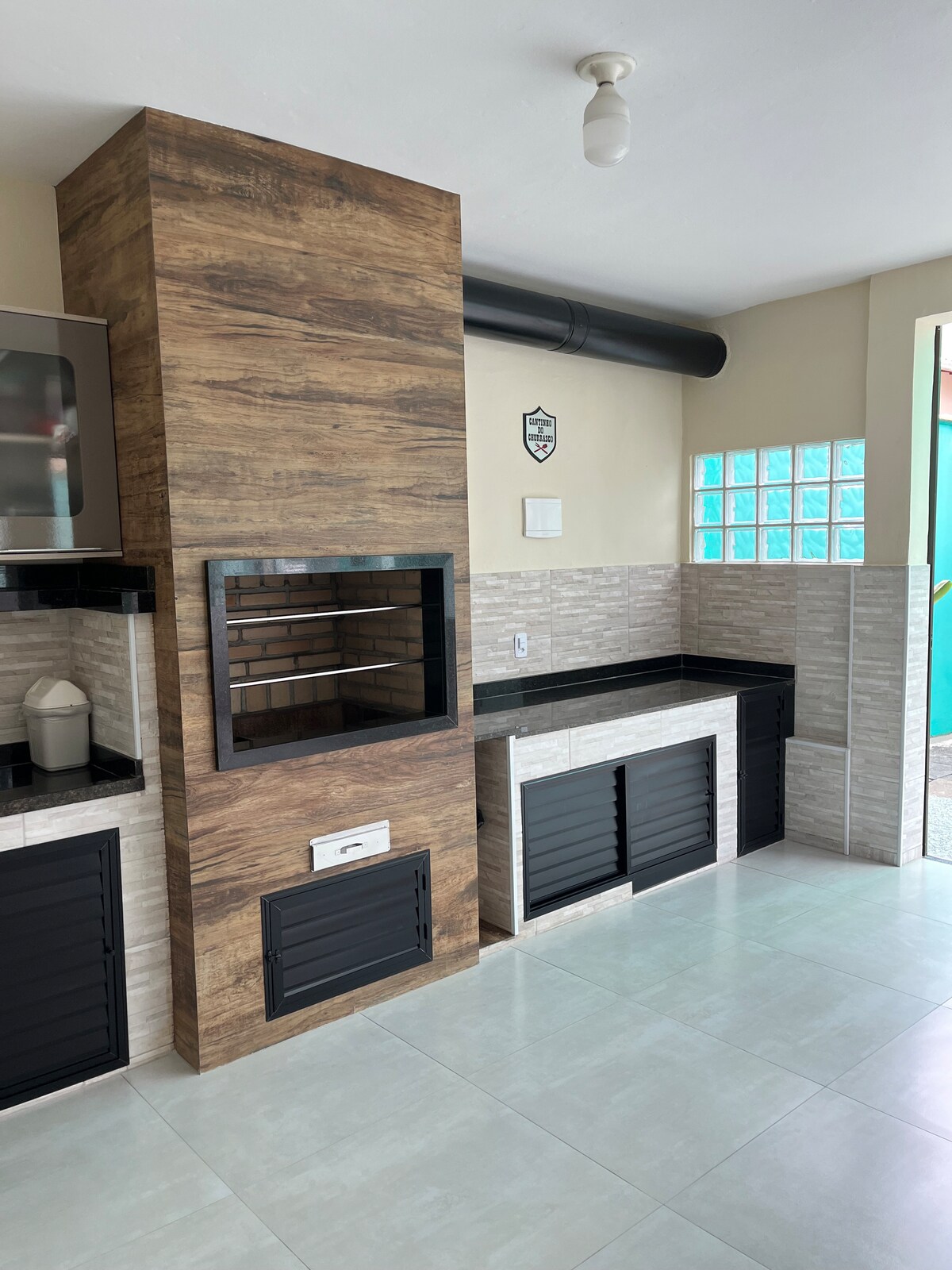 A spacious area features a modern grill integrated into a wooden panel wall. Adjacent is a countertop with sleek black cabinetry. The design incorporates light tile flooring and a window with textured glass, offering natural light to the space.