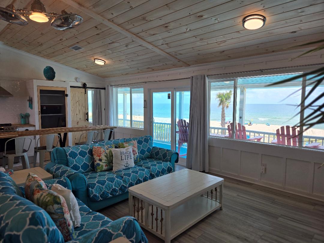 A light-filled living room features a cozy seating area with vibrant blue sofas, arranged around a coffee table. Large windows provide direct views of the ocean, with palm trees visible outside. An open kitchen space is seen in the background, enhancing the airy atmosphere.