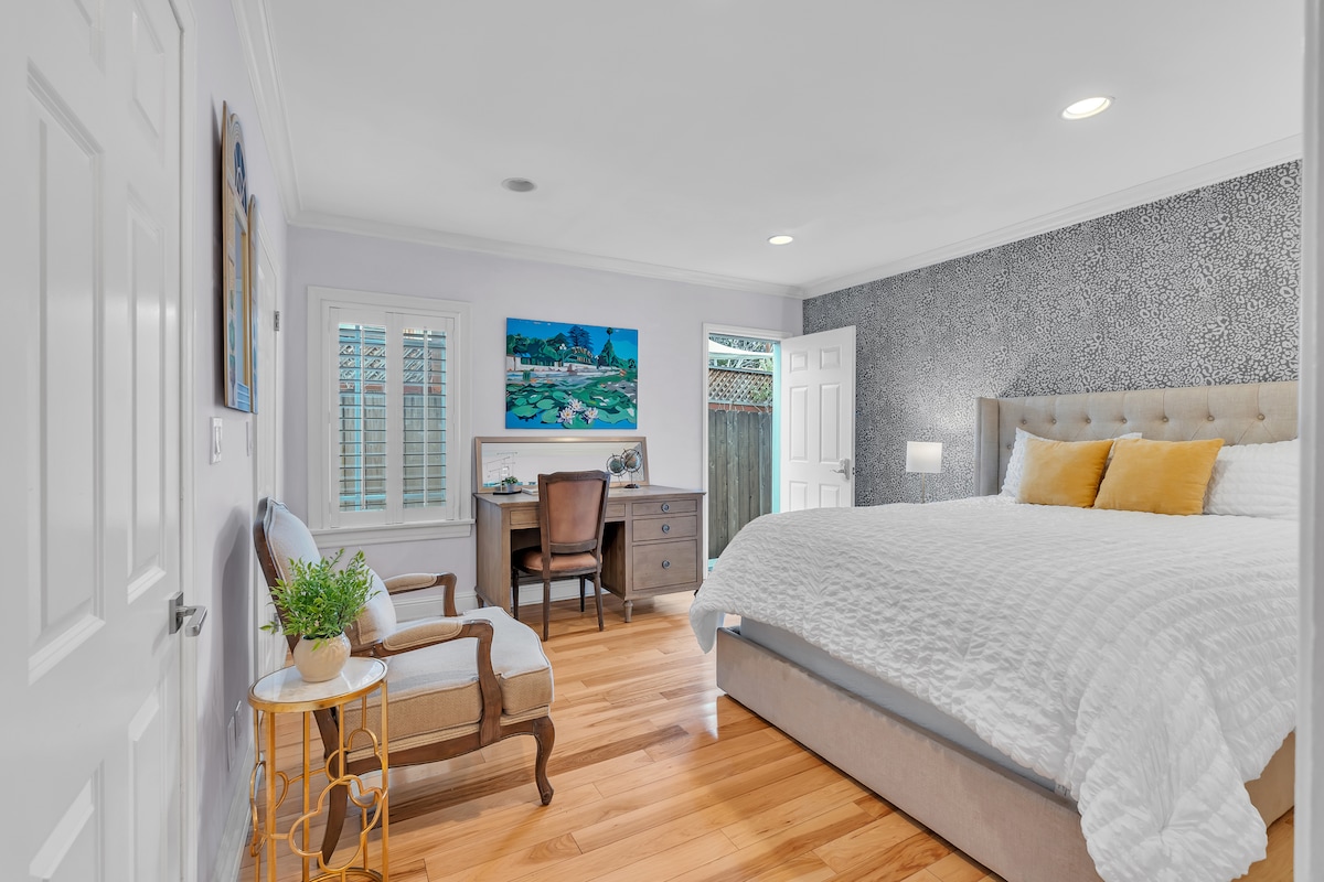 A spacious bedroom features a large bed with plush pillows and a soft quilt. A wooden desk and chair are positioned against a textured wall. Natural light enters through the window, and a small plant sits on a side table next to an armchair, creating a relaxed ambiance.