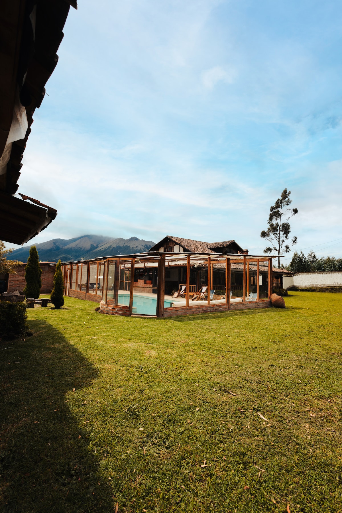 A spacious outdoor area displays a covered, heated pool surrounded by large windows. The grassy lawn is complemented by two tall trees and a view of distant mountains, adding to the serene environment. The wooden structure offers a warm and welcoming presence in the landscape.