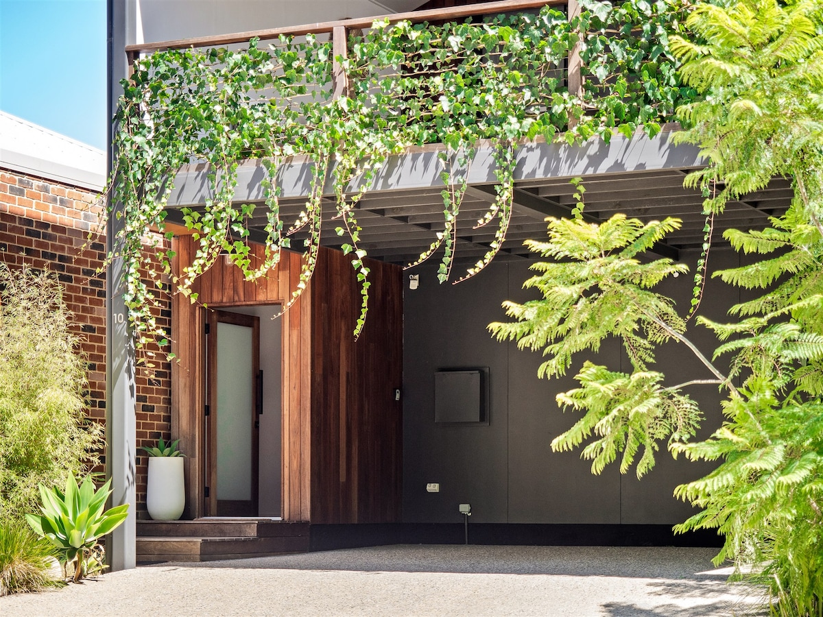 The entrance to the Leaf House is framed by lush greenery, with vines cascading from the balcony above. A wooden door leads into the home, set against a backdrop of dark walls and natural stone elements. Potted plants add to the inviting ambiance.
