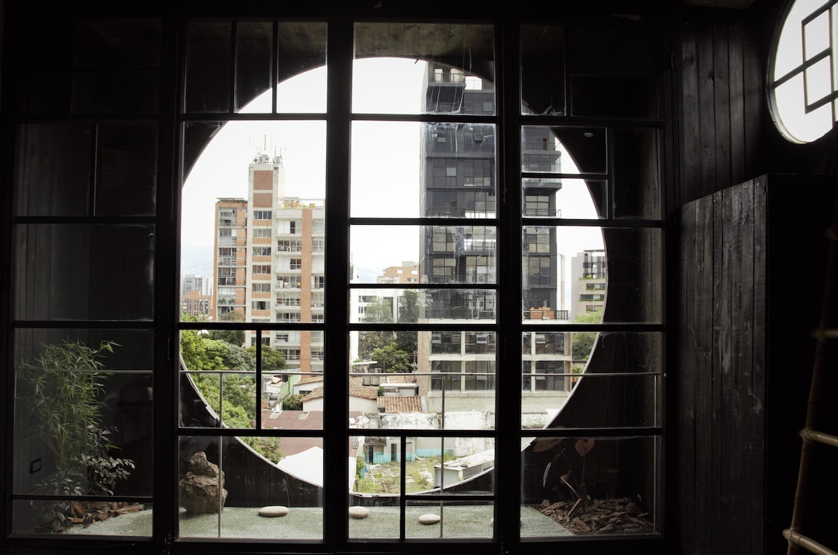 A large circular window frames an urban view, displaying a mix of buildings and greenery. The design elements include black wooden framing, creating a striking contrast with the exterior landscape. Natural light filters through, adding depth to the interior space.
