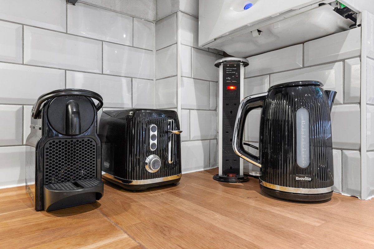A modern kitchen countertop is displayed, featuring a black coffee machine, a toaster, and a sleek electric kettle. The appliances are situated against a backdrop of glossy white subway tiles and wooden surfaces, providing a functional and stylish cooking area.