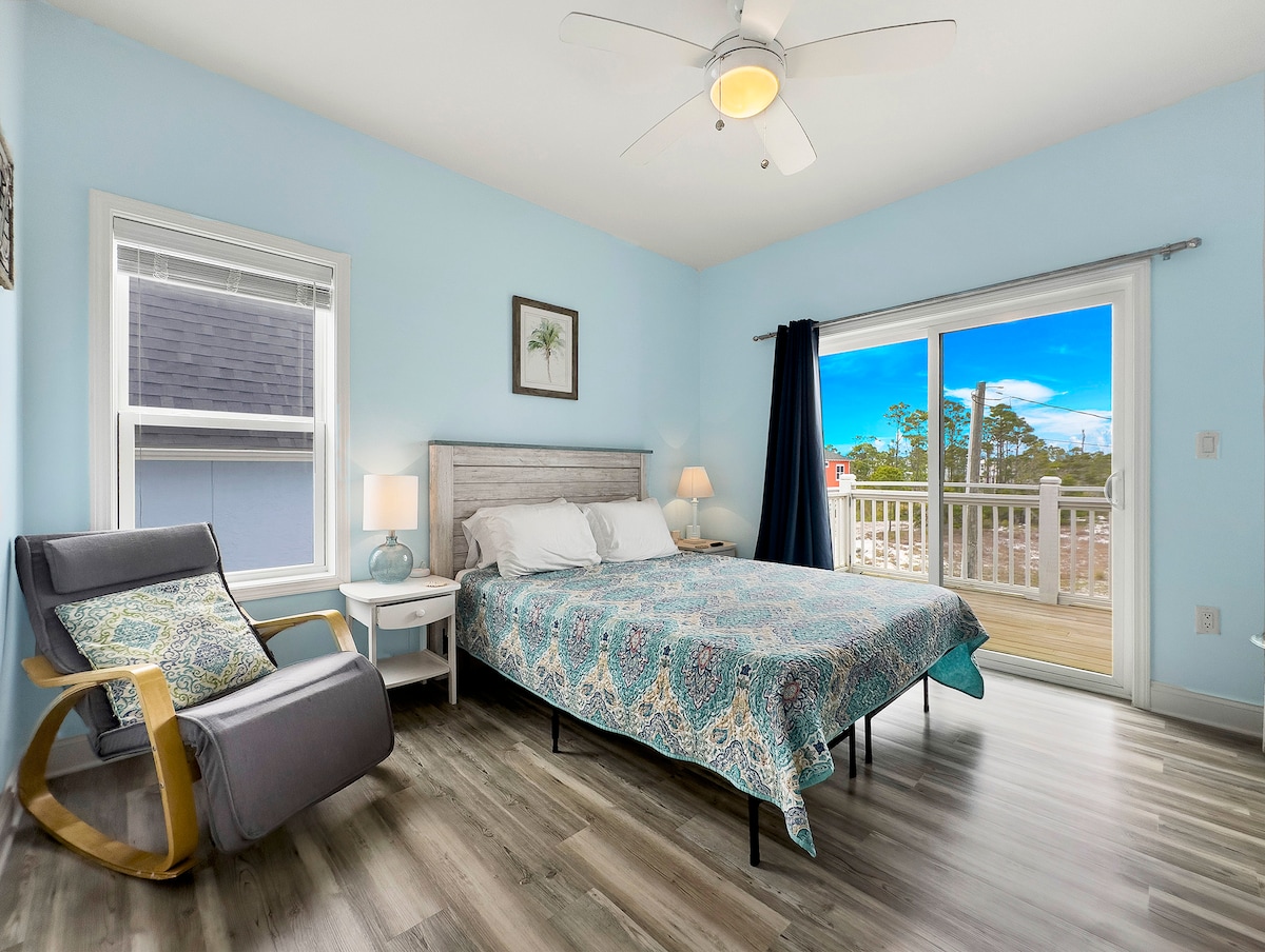 A well-lit bedroom features a spacious bed with a patterned blue coverlet. A cozy rocking chair is positioned beside a side table with a lamp. Large sliding glass doors provide access to a deck, offering outdoor views and abundant natural light.
