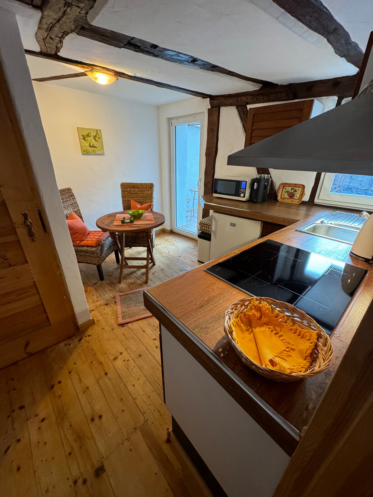 A well-equipped kitchen features wooden cabinetry and a warm wooden floor. A round table with two wicker chairs is set for relaxation, nearby a glass door providing access to the outside. Kitchen appliances, including a stovetop and microwave, are neatly arranged for convenience.