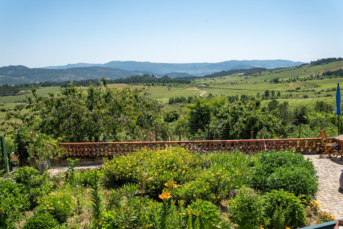 A lush garden area is visible, featuring a variety of greenery and colorful flowers. Scenic hills and valleys extend in the background, under a clear blue sky. A wooden railing borders the garden, providing a tranquil view of the surrounding landscape.