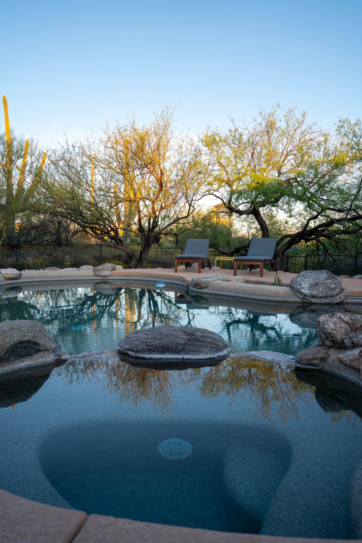A private heated pool is surrounded by natural desert landscaping, featuring large boulders and native plants. Two lounge chairs are positioned nearby, offering a place to relax. The tranquil water reflects the surrounding flora and clear blue sky.