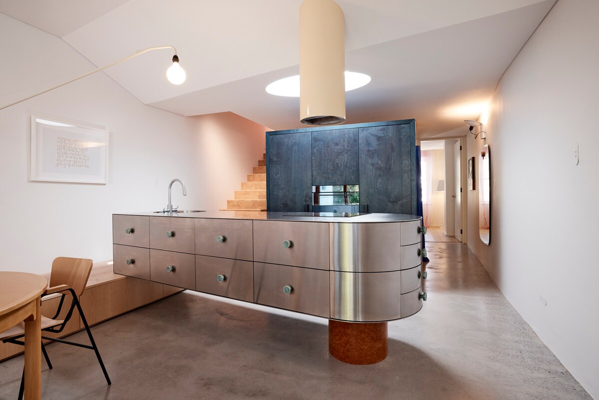A modern kitchen area features a sleek, curved stainless steel island with ample storage and stylish black cabinetry. Natural light is allowed in through a circular skylight above, illuminating polished concrete floors and complementing the minimalist design.