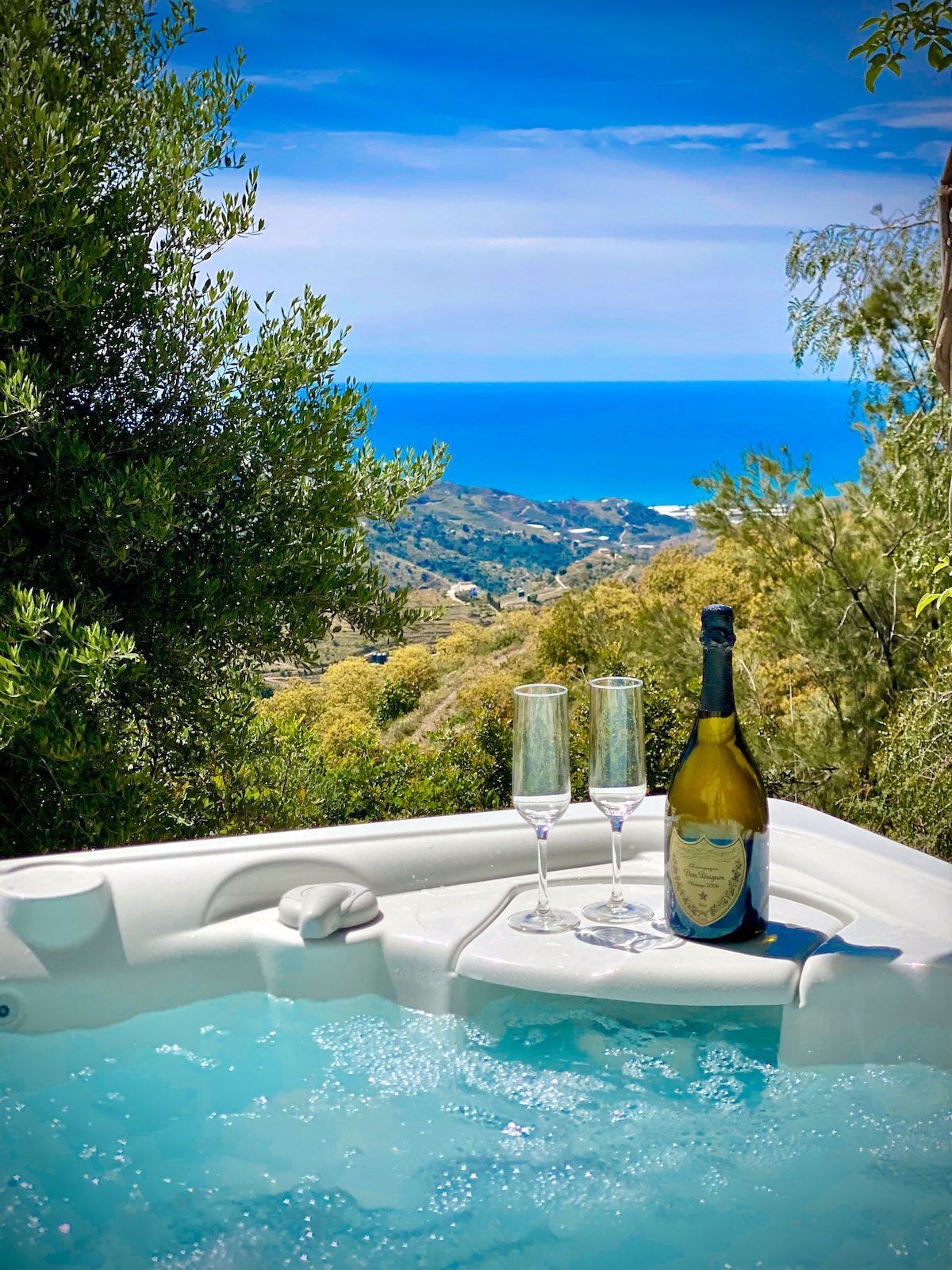 A heated spa with bubbles invites relaxation, positioned against a backdrop of rolling greenery and a panoramic view of the sea. Two champagne flutes rest beside a bottle, adding a touch of elegance to the tranquil outdoor setting.