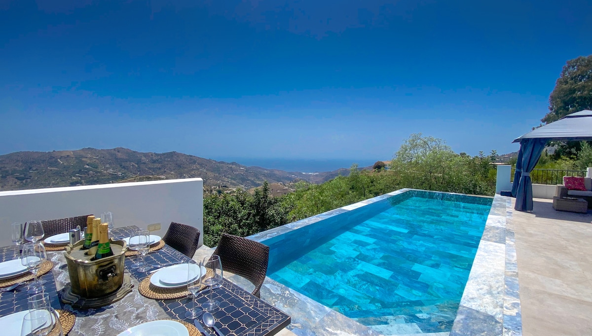 An outdoor dining area is set with an elegant table overlooking a breathtaking view of the mountains and the sea. A turquoise infinity pool extends alongside, seamlessly blending with the landscape under a clear blue sky.