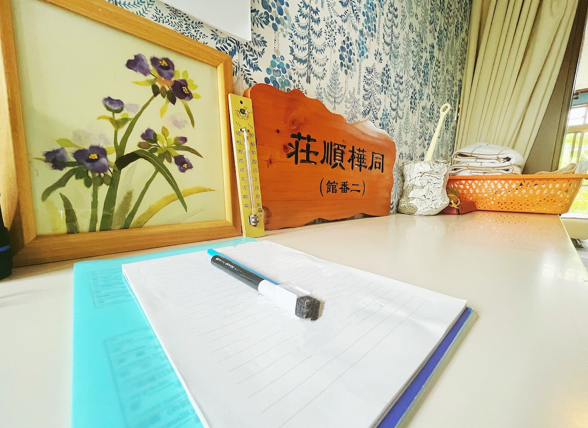 A light, wooden desk features a painting of blooming violets in a frame, alongside a decorative sign displaying the accommodation name. A lined notebook is placed on the desk, with a pen resting on top. A pastel blue folder and a woven basket are also visible.