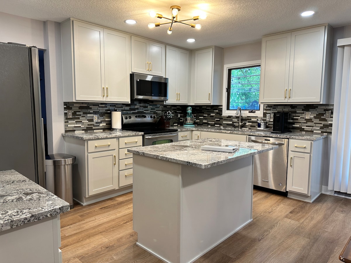 A modern kitchen is highlighted by white cabinetry and a spacious island with a gray granite countertop. Stainless steel appliances, including a microwave and oven, complement the design. A window allows natural light to illuminate the area, while a decorative backsplash adds visual interest.