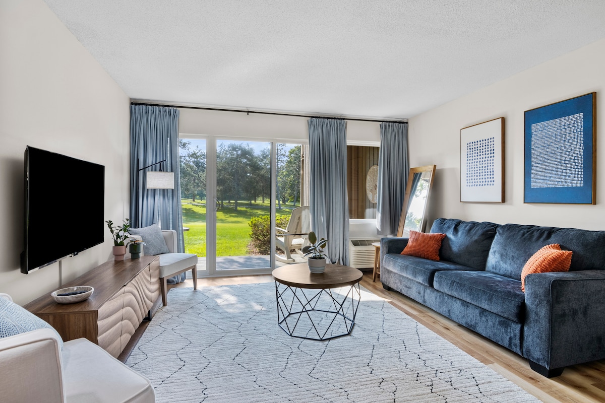 A spacious living area features a comfortable blue sofa with an orange accent pillow. A round coffee table is centered on a light area rug. Large glass doors provide access to an outdoor view of green space, complemented by tall curtains that frame the windows.