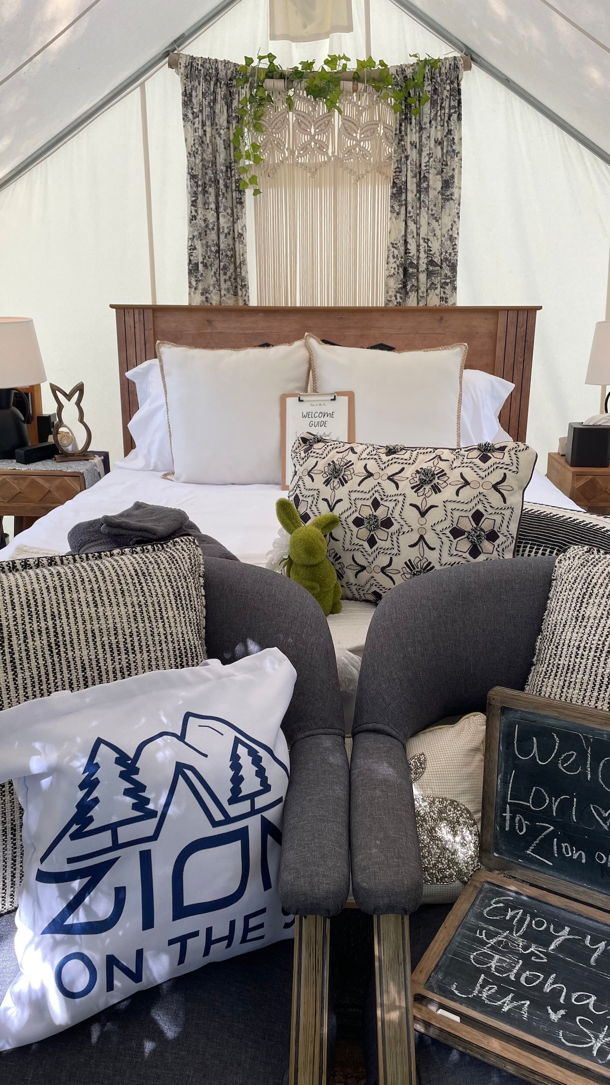 A cozy sleeping area is highlighted by a spacious bed adorned with decorative pillows and soft linens. A welcoming message is displayed on a chalkboard, while complementary armchairs and accent decor add to the warm ambiance of the tent.