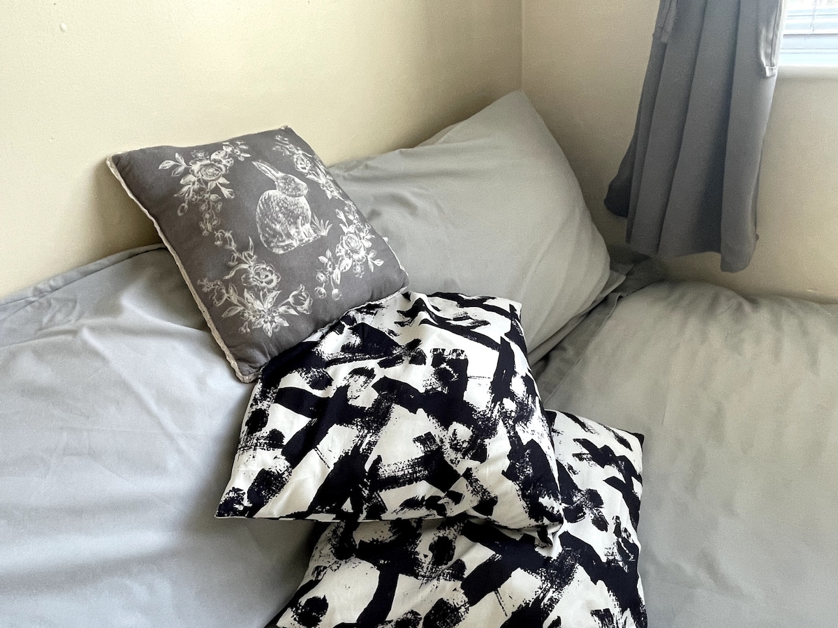 A cozy sleeping area presents a spacious bed covered with soft, light grey linens. Decorative pillows in contrasting patterns, including a floral design and abstract shapes, are arranged at the head of the bed. Natural light filters through a nearby window, casting a gentle glow.