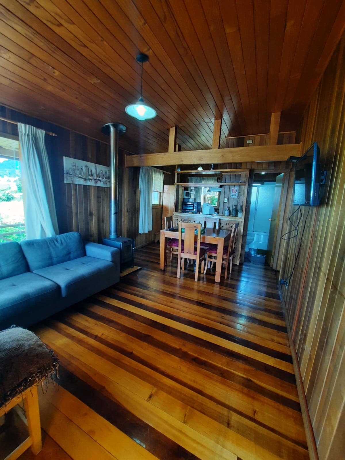 A living space features warm wooden paneling and a mix of dark and light hardwood flooring. A spacious blue couch is positioned near a wood-burning stove. A wooden dining table with chairs is visible, along with a television mounted on the wall. Natural light enters through a nearby window.
