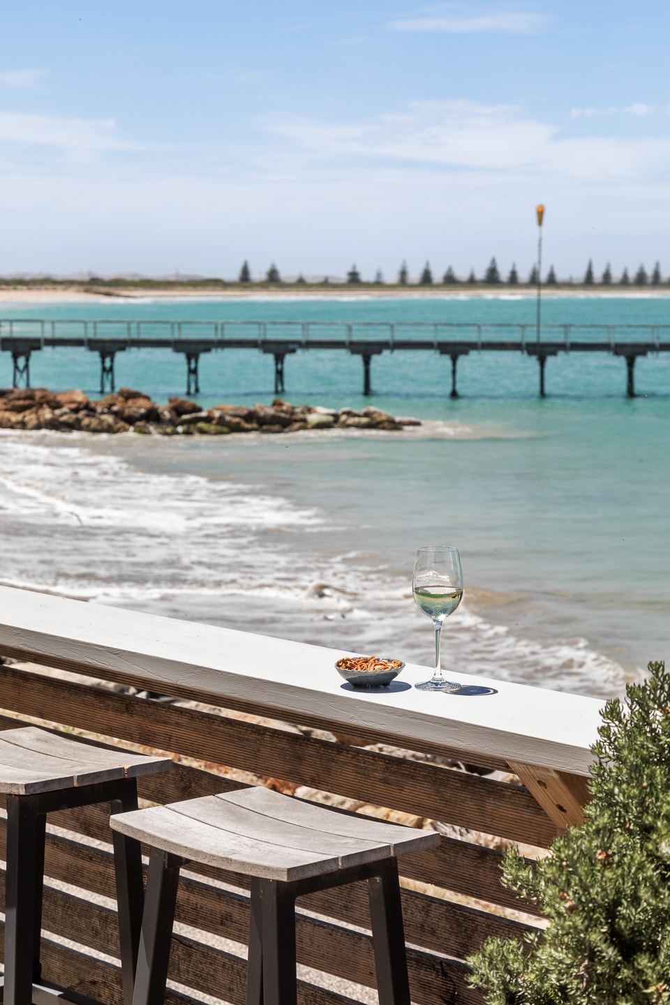 A coastal view is captured from a private deck overlooking the water. A glass of wine and a bowl of snacks are positioned on a wooden railing, with a scenic jetty stretching into the turquoise sea and gentle waves lapping at the shore.