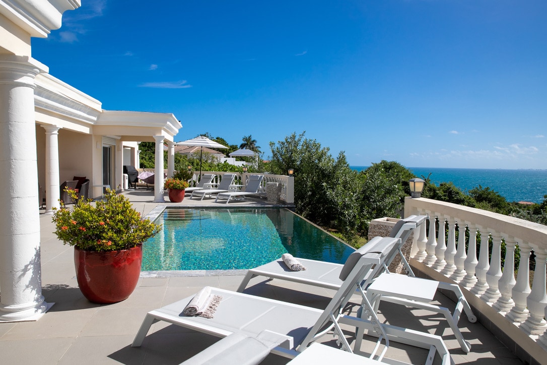 A serene pool area features a reflective swimming pool surrounded by lounge chairs. Lush greenery and tropical plants are set against a backdrop of the ocean and clear blue sky. The architectural details include white columns and a stone terrace.